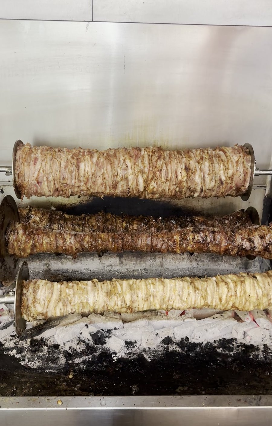 Three charcoal rotisseries loaded with marinated chicken maryland and lamb shoulder spinning above real charcoal at Yassas Greek restaurant kitchen Melbourne